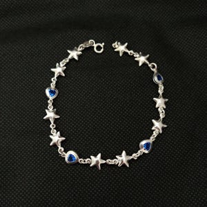 Puede incluir: Una pulsera de cadena en tono plateado que presenta una alternancia de dijes de estrellas de cinco puntas y pequeñas piedras preciosas azules en forma de lágrima. La pulsera se asegura con un cierre de resorte circular de metal pulido.