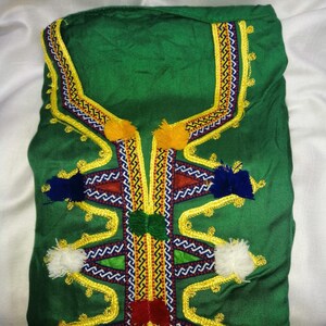 May include: A green garment with intricate embroidery featuring yellow, red, blue, and white thread. The garment has a high neckline and is adorned with colorful tassels.