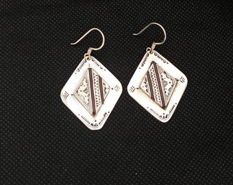 May include: A pair of silver-toned diamond-shaped earrings. Each earring features a central rectangular design in a dark reddish-brown color, surrounded by intricate patterns and a decorative border. The earrings have a hook closure.