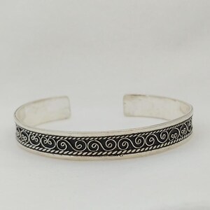 May include: A silver bangle bracelet with a black, intricate, swirl design.