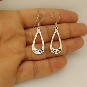 May include: A pair of silver teardrop earrings with a decorative geometric pattern at the bottom. The earrings have a simple, open design and are suspended from a hook. The earrings are displayed against a neutral background.
