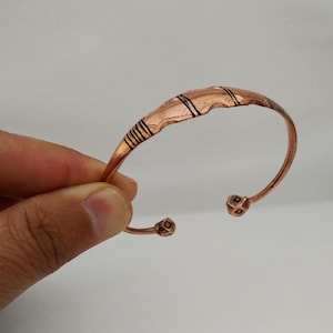 Puede incluir: Una pulsera de brazalete de color cobre con un diseño curvo. La pulsera presenta líneas negras grabadas y detalles geométricos. Los extremos de la pulsera tienen pequeños acentos en forma de cubo. La pulsera se sostiene en una mano.