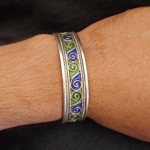 Könnte beinhalten: Silberfarbenes Armreif-Armband mit farbenfrohem Emaille-Design. Das Armband hat ein sich wiederholendes Muster aus blauen und grünen Wirbeln, das von einem gedrehten Seilmuster umrandet ist. Das Armband wird am Handgelenk einer Person getragen.