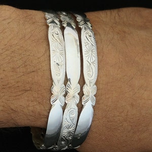 May include: Three silver bracelets with intricate floral designs. The bracelets are stacked on top of each other on a person's wrist.