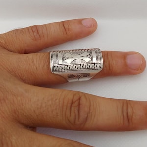 May include: A wide silver ring featuring an engraved geometric pattern across its surface. The design includes a central cross-like motif flanked by linear borders and a series of small rectangular shapes along the top and bottom edges.
