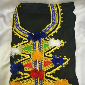 May include: Black garment with intricate embroidery featuring yellow, blue, red, and white pom-poms. The neckline is adorned with a decorative trim in blue and yellow.