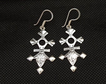 May include: A pair of silver-colored drop earrings featuring a symmetrical, geometric cross-like design with intricate etched patterns, a central circular cutout, and a pointed bottom, suspended from curved silver-colored ear wires.
