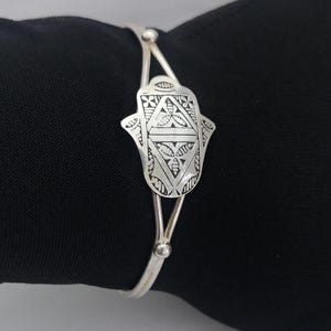 May include: A silver bracelet with a hamsa hand charm. The charm is intricately detailed with geometric patterns.