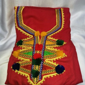May include: A red garment with intricate embroidery featuring yellow, green, blue, and red thread. The garment has a neckline with a decorative design and is adorned with pom-poms.