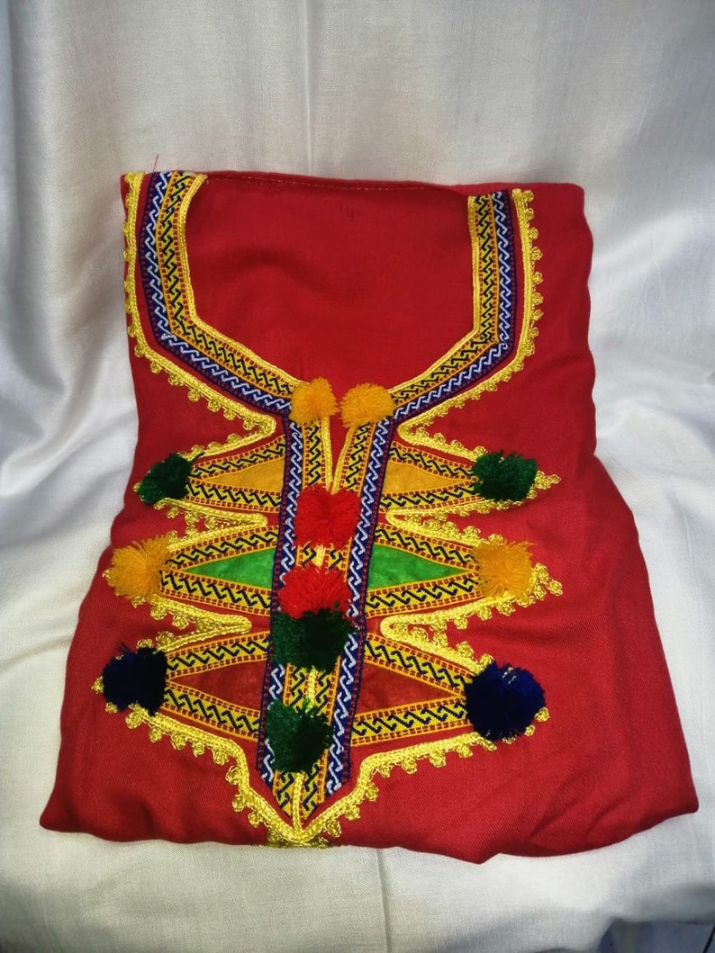 May include: A red garment with intricate embroidery featuring yellow, green, blue, and red thread. The garment has a neckline with a decorative design and is adorned with pom-poms.