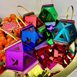 May include: A collection of colorful, geometric lanterns in shades of green, red, blue, and purple, each with a gold handle. The lanterns are arranged on a gold tray with orange flowers and a pink lotus.