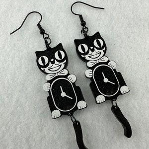 May include: A pair of black cat-shaped earrings with white accents. Each earring features a smiling cat face, a bow tie, and a clock design. The earrings have black fish hook ear wires and a dangling tail.