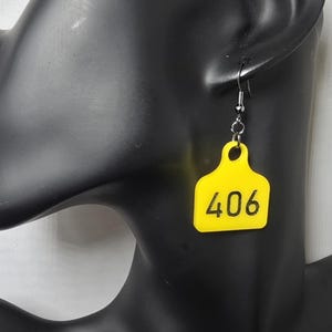 May include: A pair of yellow earrings shaped like livestock tags, each with the number 406 in black. The earrings hang from silver-colored fish hook ear wires. The earrings are displayed on a black mannequin head.
