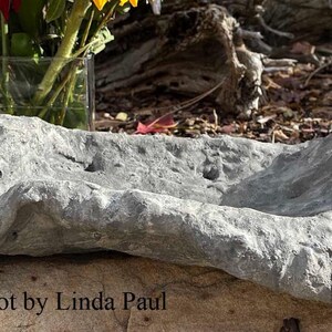 May include: A gray stone bonsai pot with a rough, textured surface. The pot is shaped like a shallow bowl with a slightly curved edge. The text "bonsai pot by Linda Paul" is visible below the pot.