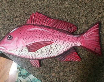 May include: A hand-painted wooden fish sculpture with pink and white scales. The fish has a red dorsal fin and tail fin.
