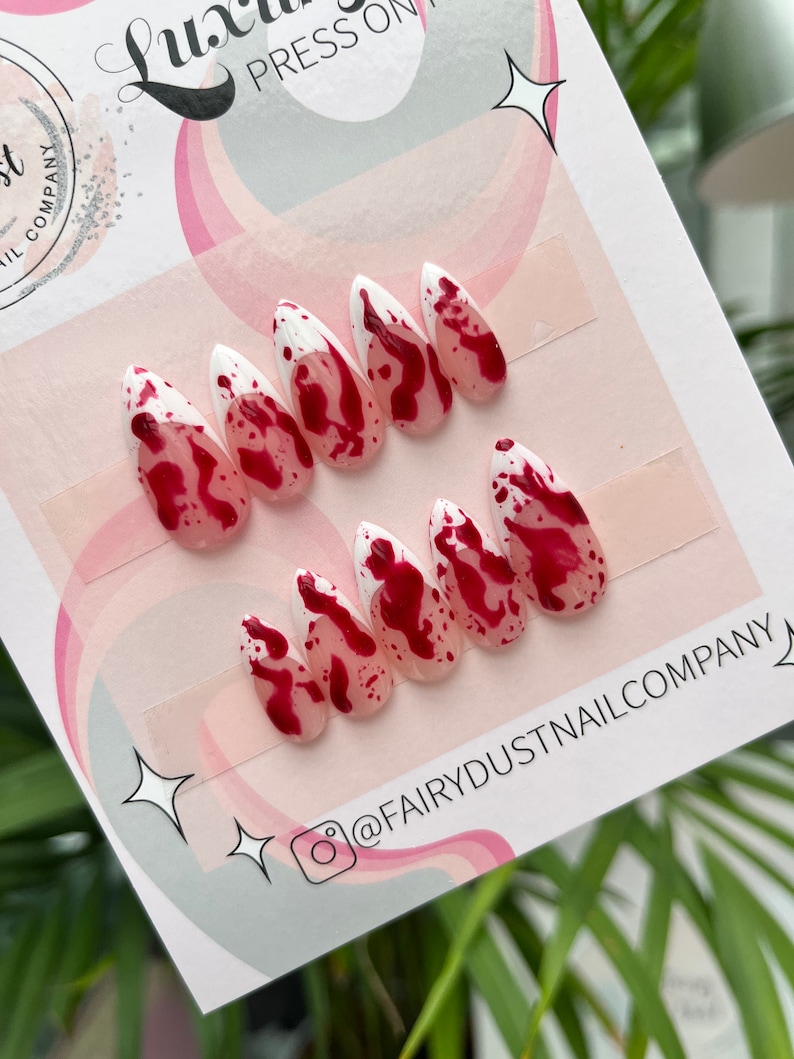 May include: Set of ten fake nails with a white tip and red blood splatter design. The nails are on a white card with the text "@FAIRYDUSTNAILCOMPANY" printed on it.