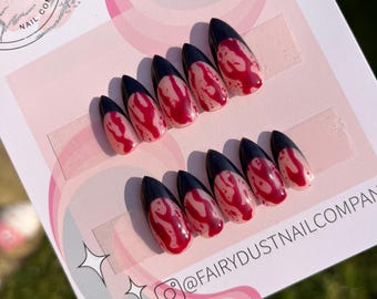 Pode incluir: Conjunto de unhas postiças com um design preto e vermelho. As unhas são longas e pontiagudas, com uma ponta preta e uma base vermelha. A base vermelha tem um efeito de sangue pingando. As unhas estão em um cartão branco com o texto "Fairy Dust Nail Company" e "@FairyDustNailCompany".