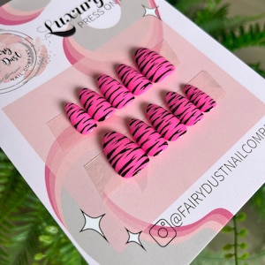May include: Pink press-on nails with black tiger stripes. The nails are arranged on a pink and white card with the text "Luxury Press On Nails" and the logo of "Fairy Dust Nail Company". The Instagram handle @FAIRYDUSTNAILCOM is also visible.
