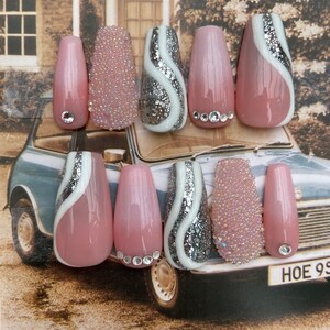 Peut inclure: Ensemble de dix faux ongles roses et argentés avec différents motifs. Certains ongles présentent un motif de tourbillon blanc avec des paillettes argentées, d'autres un motif de perle rose et blanc, et d'autres un seul strass.