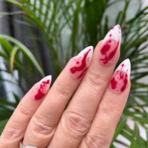 May include: A hand with white and red nail polish with a blood splatter design. The nails are long and pointed.