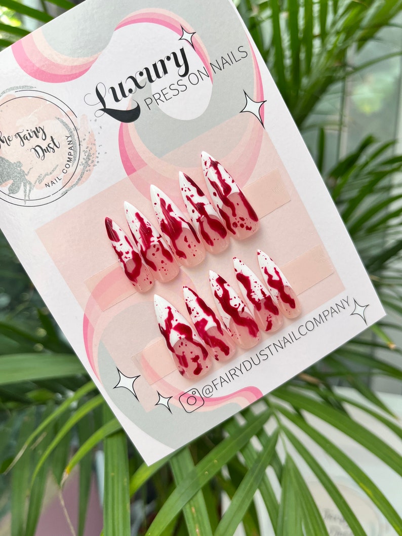 May include: A set of ten white press-on nails with a red and white blood splatter design. The nails are long and pointed, and the design is reminiscent of a horror movie. The nails are packaged on a white card with the text "Luxury Press On Nails" and "@FairyDustNailCompany".