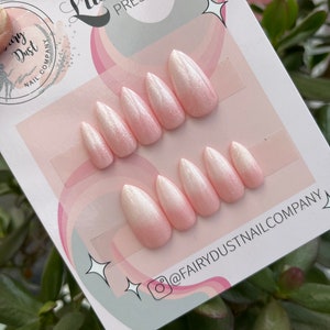 May include: A set of 10 pink and white ombre press-on nails with a glitter finish. The nails are arranged on a white card with the text "Luxury Press On Nails" and "@FairyDustNailCompany".
