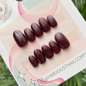 Peut inclure: Ensemble de dix faux ongles ovales de couleur bordeaux avec des paillettes argentées. Les ongles sont disposés en deux rangées sur une carte blanche avec des volutes roses et blanches. La carte porte également le texte "Luxury Press On" et "@FairyDustNailCo".