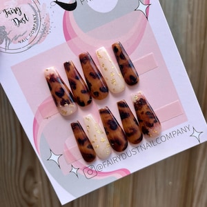 Puede incluir: Conjunto de diez uñas postizas con diseño de estampado de leopardo y detalles dorados. Las uñas se muestran sobre un fondo rosa y blanco con el texto "Luxury Press On Nails" y "@FairyDustNailCompany".