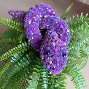 May include: A purple crocheted snake toy with colourful flecks and large blue eyes. The snake is coiled on green fern leaves. A handmade, whimsical decorative item.