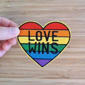 May include: A rainbow heart-shaped patch with the text "LOVE WINS" in black lettering.