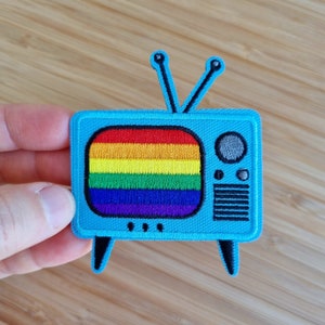 May include: A blue embroidered patch of a retro television with a rainbow screen. The television has a knob and a speaker.