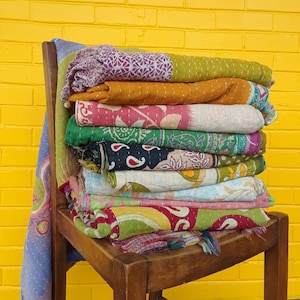 Puede incluir: Una pila de colchas kantha coloridas, hechas de saris reciclados, se exhiben en una silla de madera contra una pared de ladrillo amarillo. Las colchas están superpuestas en una variedad de patrones y colores, incluyendo rosa, verde, azul y naranja.