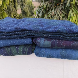 Puede incluir: Una pila de edredones teñidos con índigo con costuras blancas. Los edredones presentan varios patrones en tonos de azul, verde y morado. Los edredones están doblados cuidadosamente, mostrando los diferentes diseños y texturas.