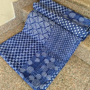 May include: A folded textile featuring a patchwork design of various blue and white patterns, including circles, concentric rings, and fan-like shapes. The fabric displays a consistent indigo-toned colour palette with a repeating block-printed aesthetic.