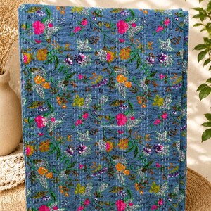 May include: A folded blue quilt with a floral pattern. The quilt features a repeating design of pink, orange, yellow, and purple flowers with green leaves. The quilt is stitched with white thread.