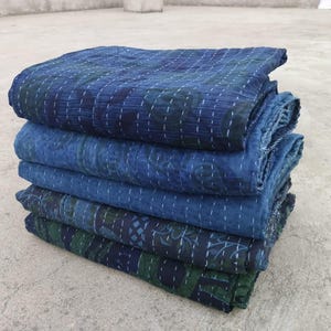 May include: A stack of five indigo blue kantha quilts with intricate stitching and floral patterns. The quilts are folded and layered on a grey concrete surface.