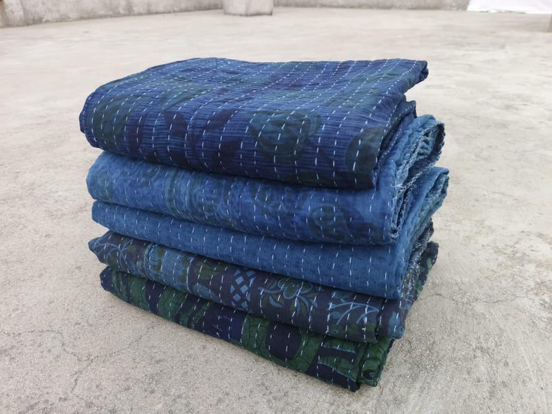 May include: A stack of five indigo blue kantha quilts with intricate stitching and floral patterns. The quilts are folded and layered on a grey concrete surface.
