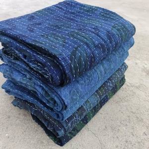 May include: A stack of four indigo blue kantha quilts with intricate stitching and patterns. The quilts are folded and stacked on a light grey concrete surface.