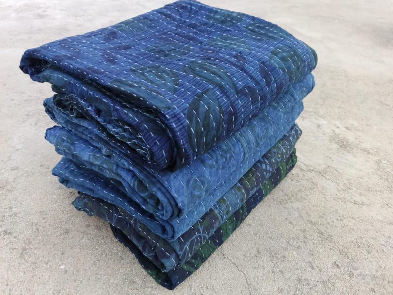 May include: A stack of four indigo blue kantha quilts with intricate stitching and patterns. The quilts are folded and stacked on a light grey concrete surface.