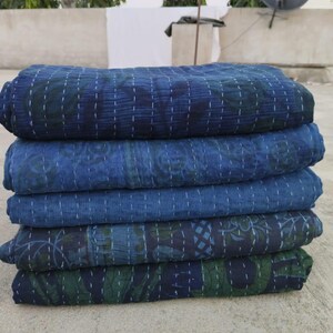 May include: A stack of five indigo blue kantha quilts with intricate floral patterns. The quilts are folded and stacked on a concrete surface.