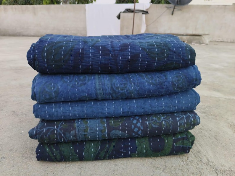 May include: A stack of five indigo blue kantha quilts with intricate floral patterns. The quilts are folded and stacked on a concrete surface.