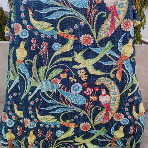 Op de afbeelding: Een blauwe kantha-deken met een bloemen- en vogelpatroon. De deken heeft een verscheidenheid aan kleuren, waaronder geel, rood, groen en blauw. De deken is gemaakt van katoen en is met de hand gestikt.