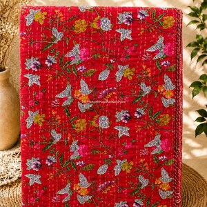 May include: A folded red quilt with a floral pattern. The quilt features a repeating design of flowers and leaves in shades of pink, yellow, and white. The quilt is stitched with white thread.