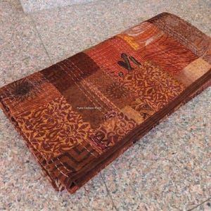 Pode incluir: Têxtil de patchwork dobrado em tons de castanho, laranja e ferrugem. O tecido é composto por vários quadrados estampados, costurados com fio branco visível. O têxtil tem uma borda decorativa e é cuidadosamente dobrado.