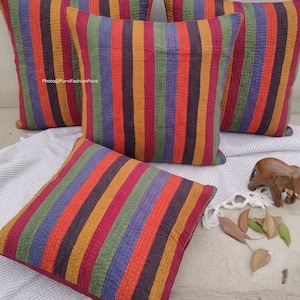 May include: Four square throw pillows with vertical stripes in shades of red, orange, yellow, green, and purple. The pillows are arranged on a white and gray patterned surface, with a small wooden elephant figurine and dried leaves nearby.