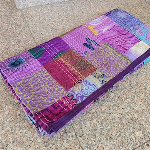 May include: A folded, patchwork quilt in shades of purple, pink, and magenta. The quilt is made of various fabric squares, each with unique patterns and textures. White stitching outlines the squares and edges.