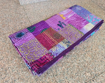 May include: A folded patchwork quilt in shades of purple, pink, and magenta. The quilt is made of various fabric squares, each with unique patterns and textures. White stitching outlines the squares and edges.