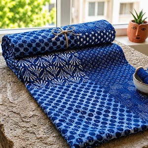 May include: A folded blue and white quilt featuring various block-printed patterns including circles, fans, and geometric shapes. The fabric is tied with a thin jute string. Next to the quilt sits a small ceramic bowl containing three spools of thread.