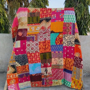 May include: A colorful patchwork quilt made from repurposed fabric scraps. The quilt is made up of squares of different colors and patterns, including floral, geometric, and paisley designs. The quilt is folded and displayed on a concrete surface.