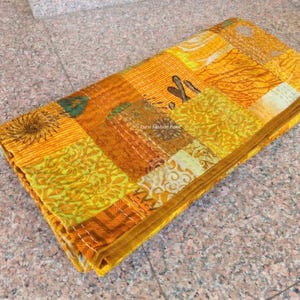 May include: Folded patchwork textile in shades of yellow, orange, and green. The fabric is composed of various patterned squares, stitched together with visible white thread. The edges are finished with a solid orange border. The textile is folded neatly.